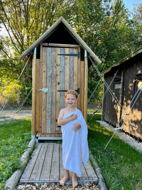 A smiling child wrapped in a white towel stands in front of a wooden hut at a sunny holiday park.