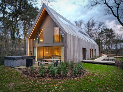Modern lodge with jacuzzi, large windows, and wood exterior, set in a tranquil wooded landscape at dusk.