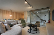 Moderne lodge woonkamer met lichte houten wanden, grote bank, eetgedeelte, en trap achter glas.