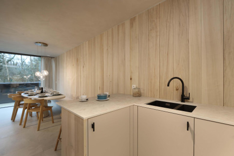 Cocina de albergue moderna con paneles de madera clara, grifo negro, comedor y ventana grande al bosque.