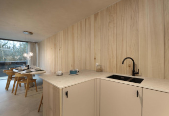 Cocina de albergue moderna con paneles de madera clara, grifo negro, comedor y ventana grande al bosque.