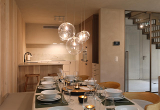 Salle à manger moderne dans un lodge avec table dressée, suspensions en verre, cuisine et escalier.