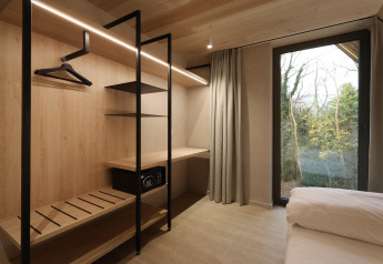 Modern lodge room with open closet, safe, desk, large window and view of the surrounding trees outside.