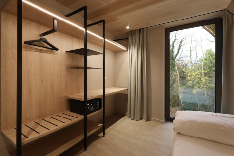 Habitación moderna de lodge con armario abierto, caja fuerte, escritorio, gran ventana y vista al bosque.