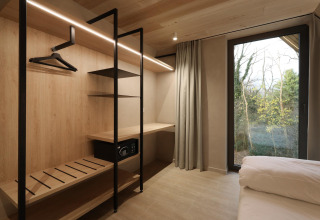 Chambre de lodge moderne avec armoire ouverte, coffre-fort, bureau, grande fenêtre et vue sur la forêt.