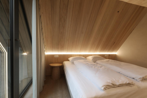 Cozy lodge bedroom with sloped wooden ceiling, two twin beds, white bedding, window, and soft LED lighting.