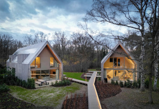 Dos cabañas modernas de madera con grandes ventanales unidas por una pasarela, ubicadas en un bosque.