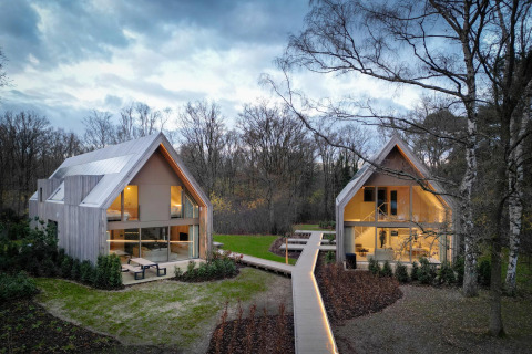 Dos cabañas modernas de madera con grandes ventanales unidas por una pasarela, ubicadas en un bosque.