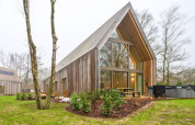 Moderne Holzlodge mit Panoramafenstern und Whirlpool, umgeben von Bäumen und Garten.