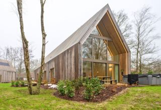 Contemporary wooden lodge with large glass windows, terrace seating, and outdoor Jacuzzi in nature.