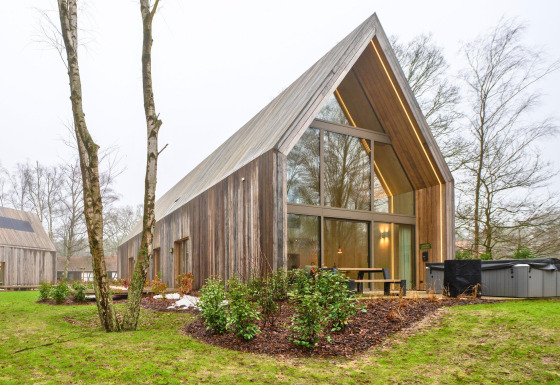 Lodge de madera moderna con grandes ventanales y jacuzzi exterior, rodeada de naturaleza y árboles.