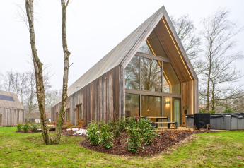 Moderne houten lodge met grote ramen, buitenterras en jacuzzi, omgeven door bomen en groen.