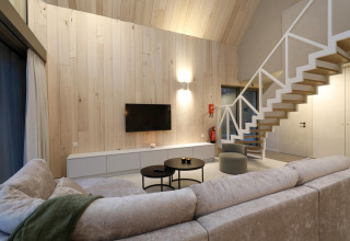 Modern lodge living room with wood paneling, staircase, gray sectional, round tables, and wall TV.