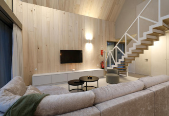 Salon de lodge moderne avec murs en bois clair, escalier, canapé gris, tables rondes et télévision.