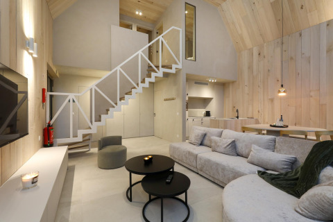 Acogedora cabaña moderna con paneles de madera clara, sofá grande, escalera abierta y cocina integrada.