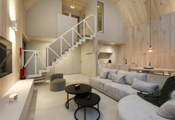 Acogedora cabaña moderna con paneles de madera clara, sofá grande, escalera abierta y cocina integrada.