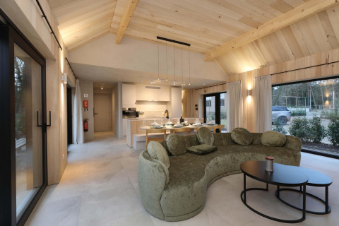 Sala moderna de Villa Lodge con sofá verde curvo, comedor y grandes ventanales en Woodz Lodges, Bélgica.