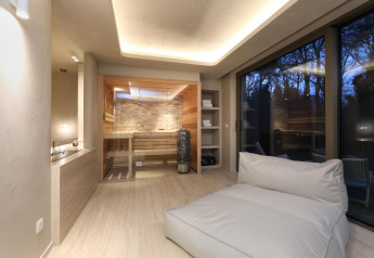 Moderne wellness-suite met houten sauna, grote loungezetel en ramen met uitzicht op de boomrijke tuin.