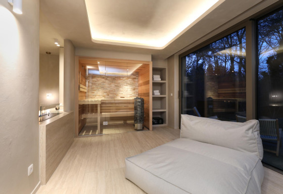 Suite de bienestar moderna con sauna de madera, sillón cómodo y grandes ventanas vistas al bosque al atardecer.