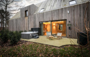 Buitenaanzicht van de Wellness Suite bij Woodz Lodges in België met jacuzzi, terrasstoelen en houten gevel.