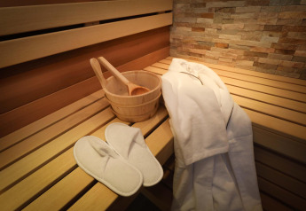 Sauna bij Wellness Suite, Woodz Lodges, België, met houten emmer, witte badjas en slippers op bank.