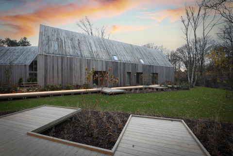 Een moderne houten lodge met schuin dak en terrassen, gelegen in een groene omgeving bij zonsondergang.