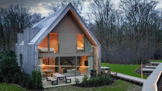 Lodge moderna con grandi finestre e rivestimento in legno immersa in una radura boschiva al tramonto.