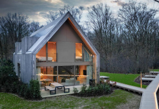 Moderne lodge met veel glas en houtbekleding in een bosrijke omgeving bij het vallen van de avond.