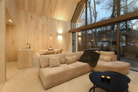 Salón moderno de una lodge con grandes ventanales, paneles de madera clara, sofá y vistas al bosque.