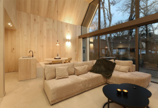 Salón moderno de una lodge con grandes ventanales, paneles de madera clara, sofá y vistas al bosque.