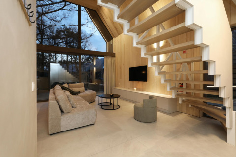 Moderne lodge woonkamer met grote ramen, houten panelen, zwevende trap en comfortabele beige zetel.