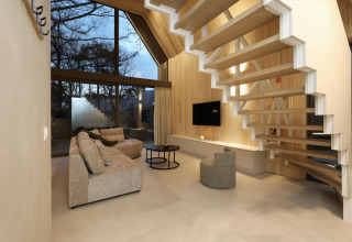 Moderne lodge woonkamer met grote ramen, houten panelen, zwevende trap en comfortabele beige zetel.