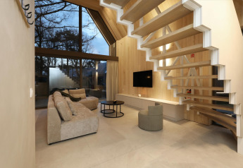 Modernes Lodge-Wohnzimmer mit großen Fenstern, Holzpaneelen, schwebender Treppe und beigen Sofas.