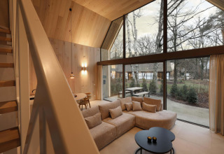 Vista interior de un lodge moderno con grandes ventanales, paredes de madera, sofá y entorno natural.