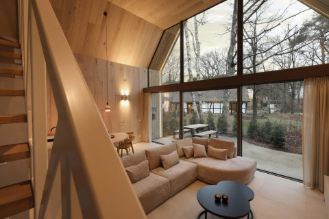 Vista interna di un lodge moderno con grandi finestre, pareti in legno, divano e vista sulla natura.