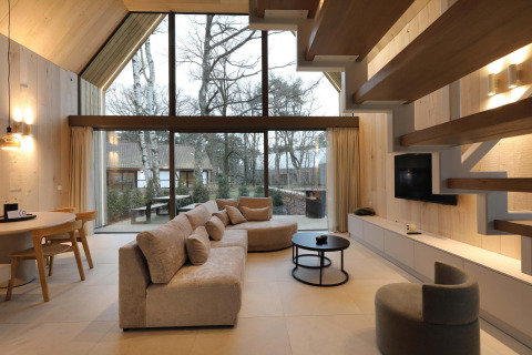 Sala de estar de lodge moderno con grandes ventanales, sofá cómodo, techo de madera y vista al bosque.
