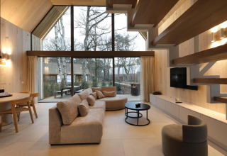 Sala de estar de lodge moderno con grandes ventanales, sofá cómodo, techo de madera y vista al bosque.