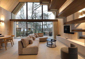 Salon de lodge moderne avec grandes fenêtres, canapé confortable, plafond en bois et vue sur la forêt.
