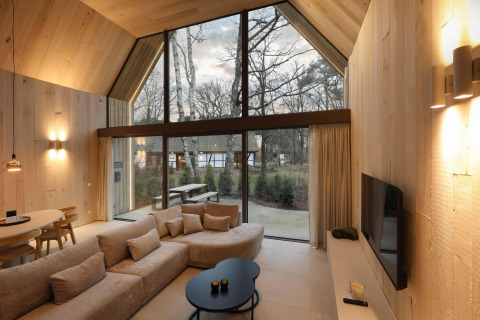 Moderne lodge met hoge ramen, lichte houten inrichting, gezellige zithoek en uitzicht op natuur.