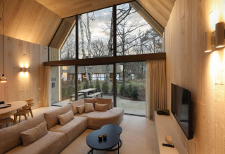 Moderne lodge met hoge ramen, lichte houten inrichting, gezellige zithoek en uitzicht op natuur.