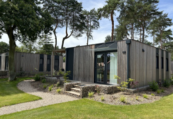 Modern chalet Domus 4 met houten gevel en grote ramen, omgeven door hoge bomen en groene natuur.