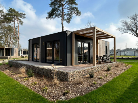 Moderne Belsano 6 lodge met houten terras en pergola, omgeven door groen en hoge bomen buiten.
