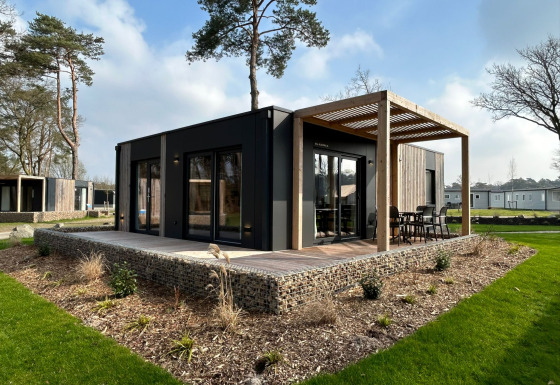 Moderne Belsano 6 lodge met houten terras en pergola, omgeven door groen en hoge bomen buiten.