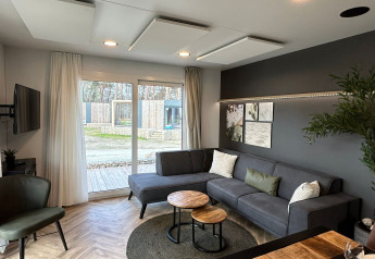 Sala moderna en la cabaña Belsano 6 en Zilverstrand, Bélgica, con sofá gris y grandes ventanales.