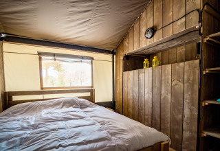Vista interna di una tenda safari accogliente con pareti di legno, letto matrimoniale e luce naturale dalla finestra.
