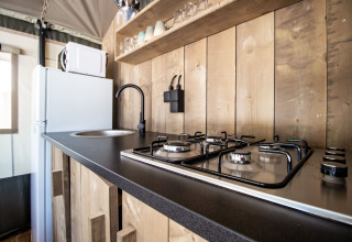 Moderne Küche in einem Safarizelt mit Gaskochfeld, Spüle, Kühlschrank und Holzregalen.