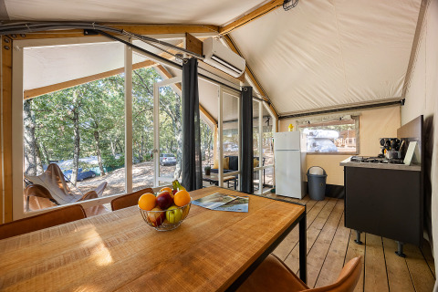 Interior of a safari tent with dining table, kitchen, fridge, and view of the forested campsite outside.