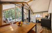 Interior of a safari tent with dining table, kitchen, fridge, and view of the forested campsite outside.