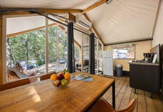 Interior de una tienda safari con mesa de comedor, cocina, nevera y vista al bosque del camping exterior.