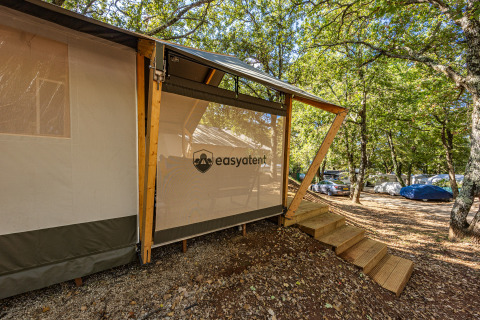 Safari-telt på campingplads med trætrappe og skov omkring under grønne trækroner, let skygge.